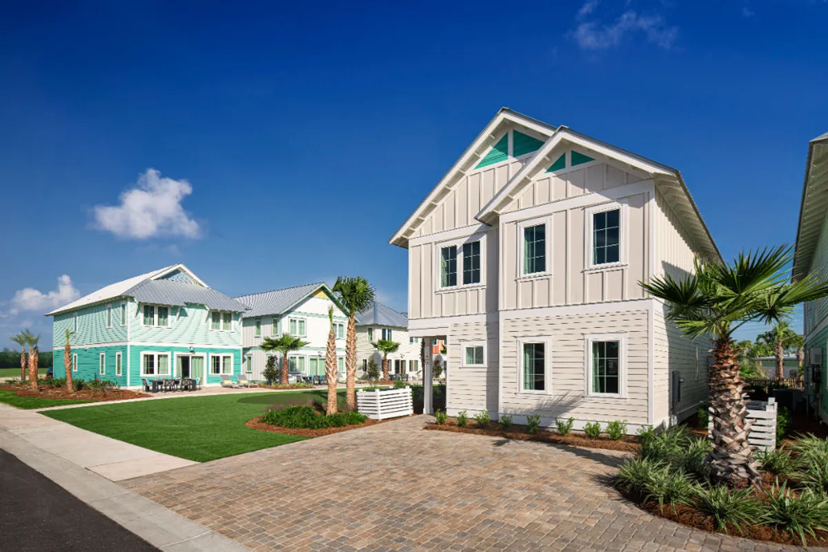 Two-story coastal-style beach cottages with pastel and neutral exteriors are shown with manicured lawns bordered by young palm trees. A wide paved driveway and walkways lead to the entrances. The setting is bright and welcoming, with a clear blue sky above.