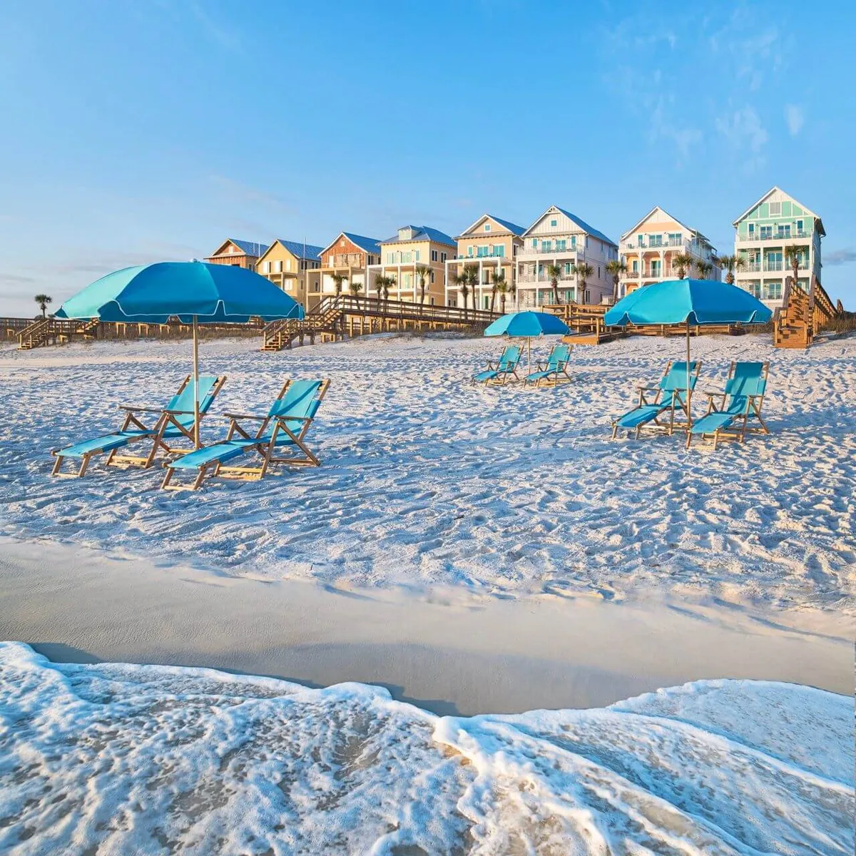 A sun-drenched row of cheerful, pastel beach cottages overlooks a pristine sandy shoreline, dotted with inviting blue umbrellas and lounge chairs. Gentle ocean waves lap the beach, while wooden boardwalks meander from the cottages directly to the surf, setting the scene for leisurely walks and carefree beach days.