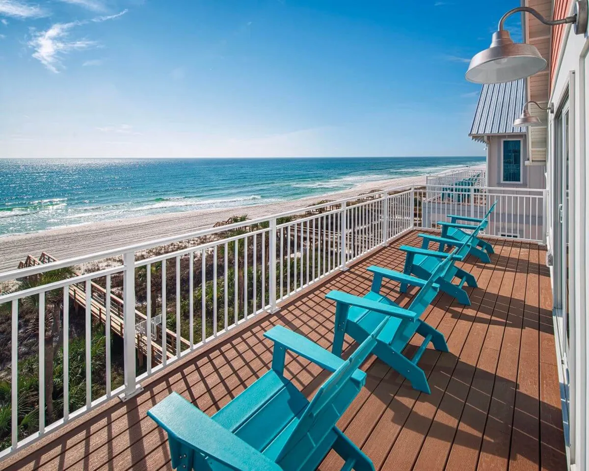 A serene balcony scene with turquoise Adirondack chairs lined up behind a white metal railing, overlooking a vast sandy beach and gentle ocean waves. A private wooden boardwalk leads from the villa to the shore, and the perspective suggests a peaceful sunrise or sunset setting, ideal for relaxation and reflection.