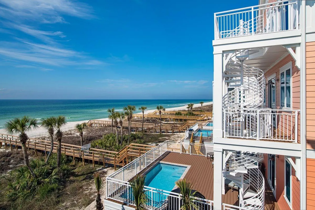 A stunning shot of a private heated pool with a spacious deck bordered by a multi-level balcony and a white spiral staircase. A wooden boardwalk leads directly onto the powdery white sands and turquoise Gulf waters, with palm trees and sand dunes framing the coastal scene. The setting exudes a blend of luxury, relaxation, and direct beach access.