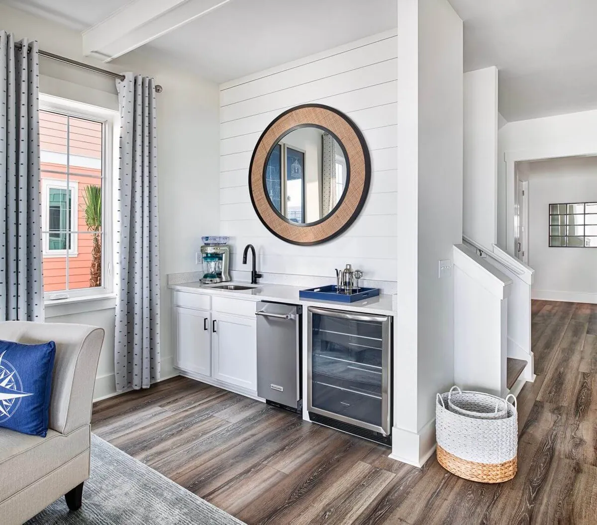 The wet bar area is artfully arranged with a sleek sink, mini fridge, and a large decorative mirror, set against a backdrop of soft natural light. The open design connects to the adjacent living space, inviting guests to gather for drinks and conversation before heading poolside or to the dining area.