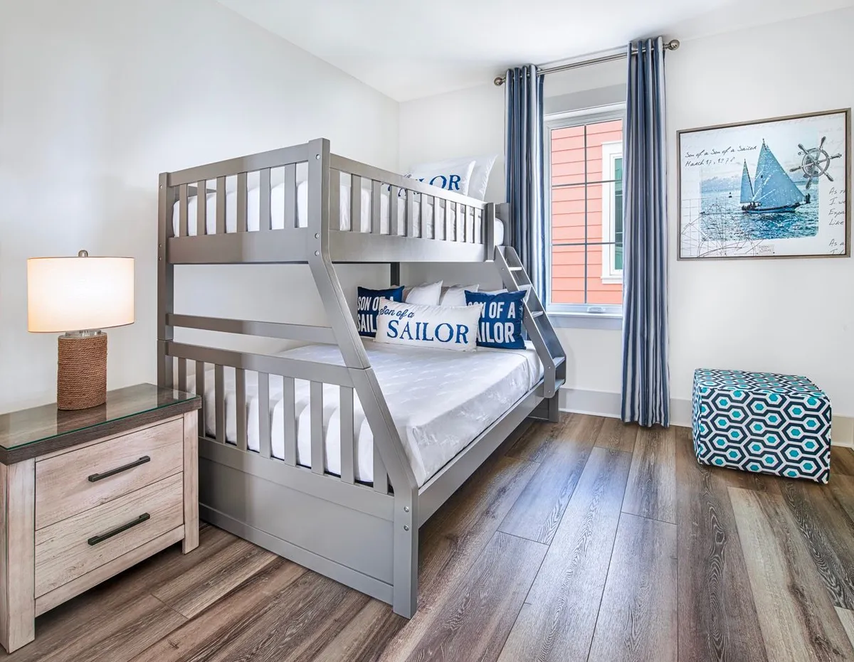A cozy, nautical-themed bunk alcove featuring a gray bunk bed with a twin over full configuration, blue and white 'Son of a Sailor' pillows, a patterned blue ottoman, and sailboat wall art. The room is inviting and clearly designed with children and families in mind, offering both comfort and playful style.