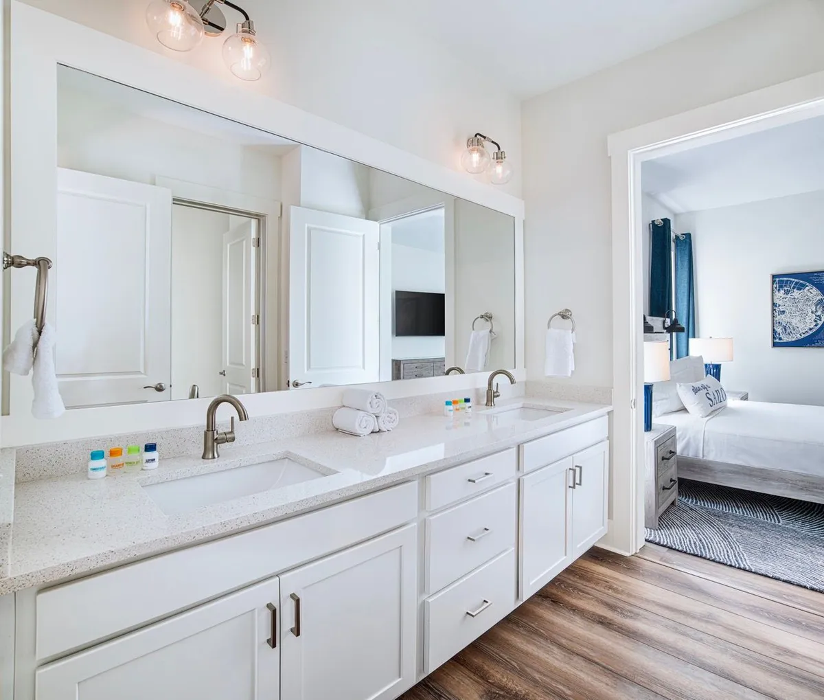 A sleek, spa-inspired bathroom with a double vanity topped with quartz, modern faucets, a large mirror, and wall-mounted light fixtures. The wood-style flooring and bright, airy feel evoke a sense of luxury and relaxation. The adjacent bedroom is visible through an open door, emphasizing the seamless connection between sleep and self-care spaces.