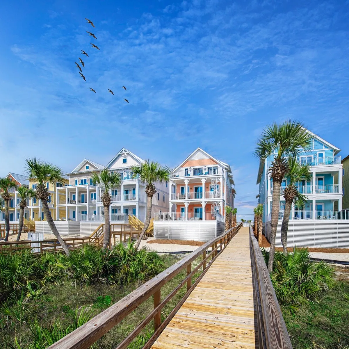 A row of colorful beachfront houses with balconies overlooks a wooden boardwalk that winds through palms toward the white sand beach. A flock of birds flies in formation overhead, enhancing the sense of a vibrant, lively coastal environment. The scene is cheerful and picturesque, inviting exploration and outdoor activities.