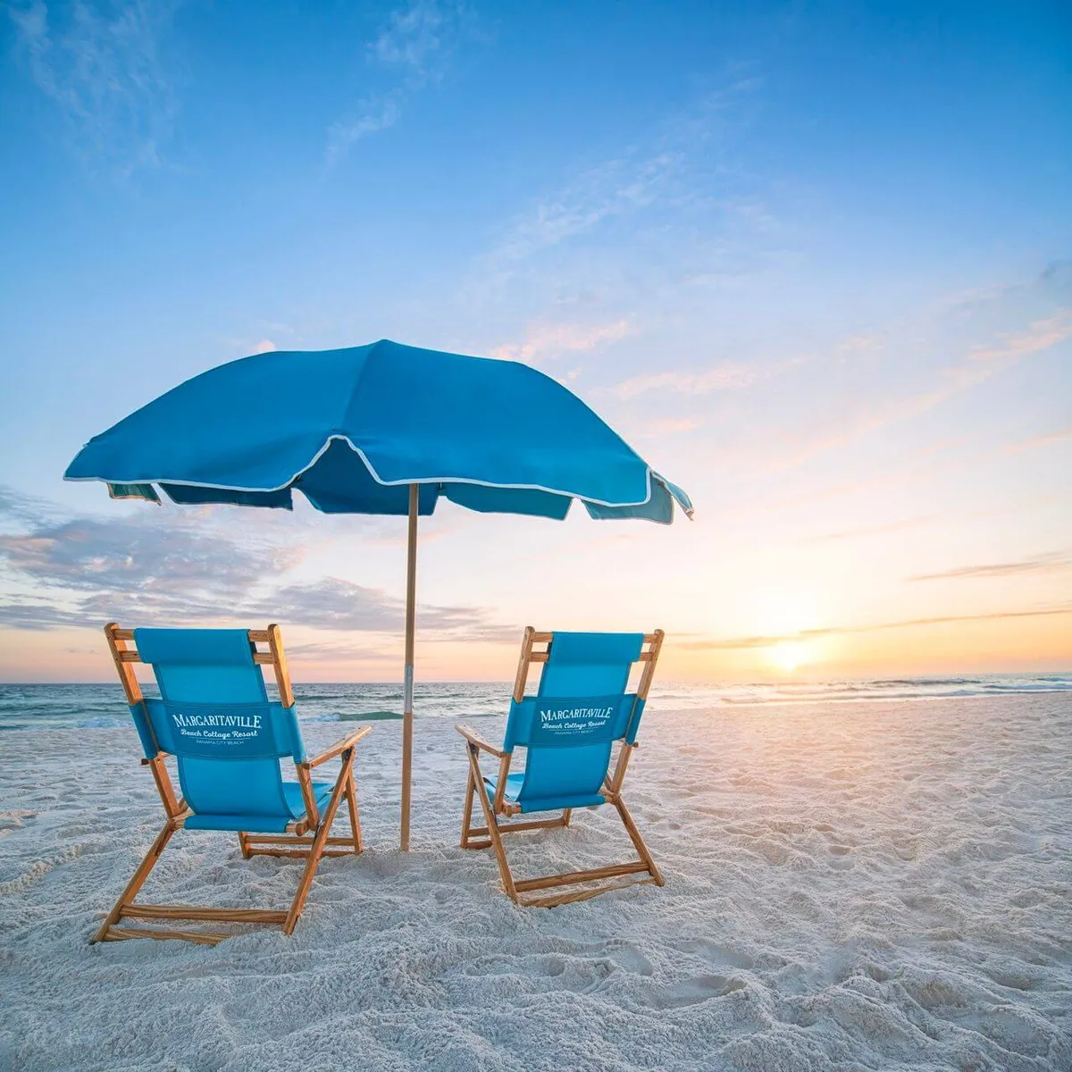 A serene beachfront scene with two blue Margaritaville-branded beach chairs under a large blue umbrella. The pristine white sand stretches towards the Gulf of Mexico, where the sun sets over the calm water, creating a tranquil and inspiring backdrop.