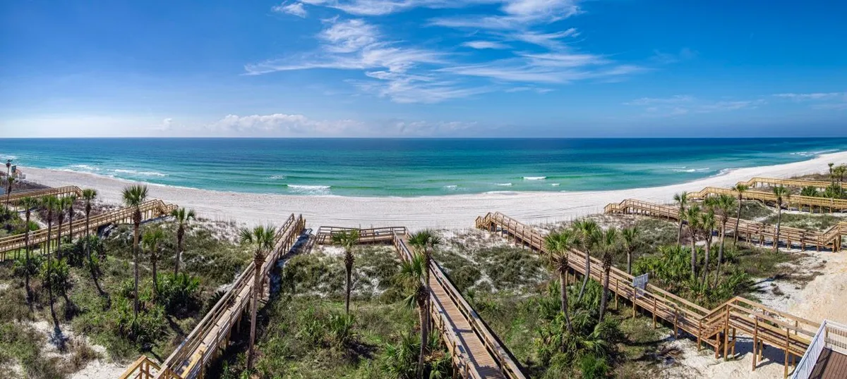 A panoramic view of private dune walkover boardwalks leading to an uncrowded, white sandy beach with calm turquoise waters. The blue sky with scattered clouds completes the tranquil, open-air environment.