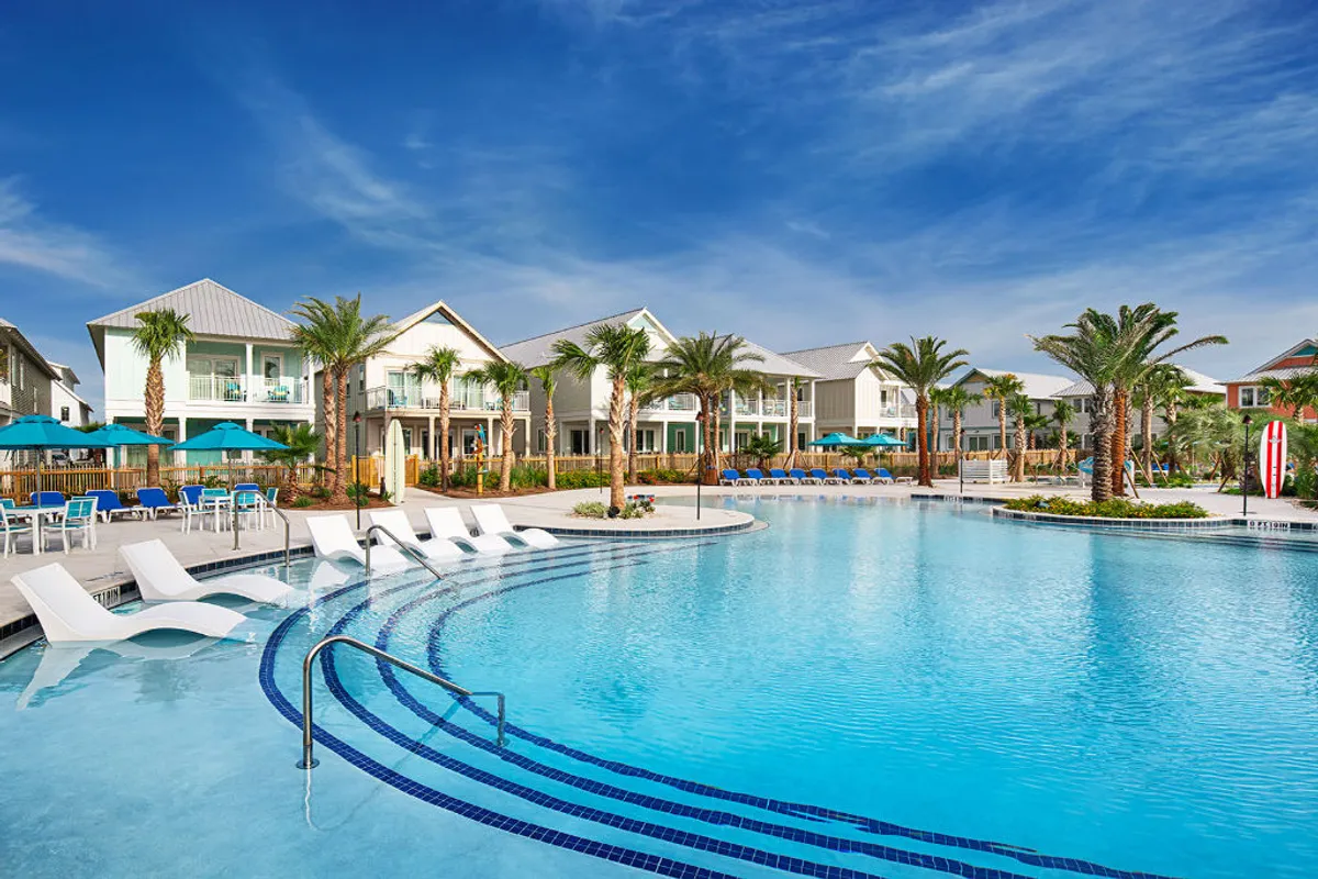 A large resort-style swimming pool sits at the heart of the property, surrounded by modern, coastal-style beachfront villas with private balconies. Tall palm trees and lush landscaping create a tranquil, tropical setting. Poolside seating features umbrellas and built-in loungers, inviting relaxation and socializing. The pool area is just steps from the beach, with glimpses of the Gulf visible beyond the villas.