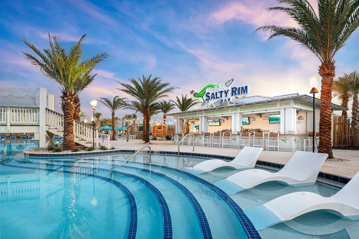 A lively poolside view with the Salty Rim Bar’s neon signage glowing above a curved swimming pool. Submerged lounge chairs, tall palm trees, and an elevated deck with seating create an inviting space for both relaxation and socializing, perfectly framed for sunset enjoyment.