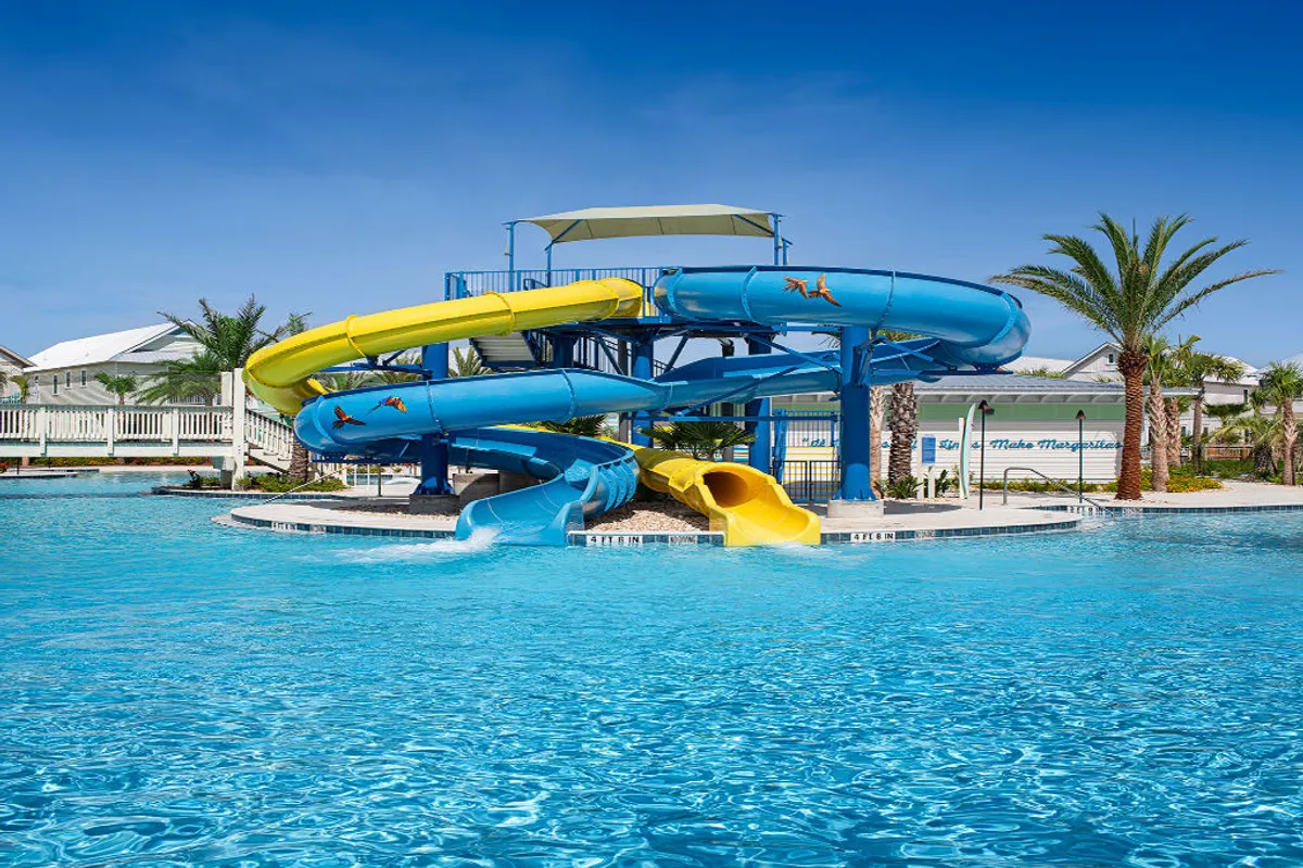 This image showcases large, colorful water slides (blue and yellow) emptying into a heated outdoor pool, surrounded by palm trees and a poolside shade canopy. The scene is lively, with space for both active fun and relaxation.