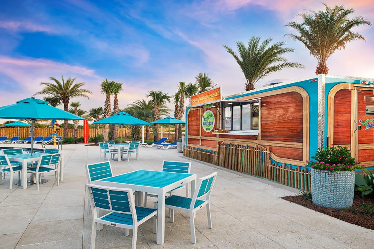 This image captures a lively outdoor dining area featuring turquoise and white tables with matching umbrellas, set against the backdrop of a retro-style wooden food and beverage kiosk and framed by tall palm trees. The scene is bathed in warm sunset hues, creating a festive and tropical atmosphere.