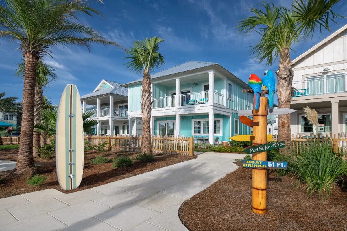 A turquoise two-story beach house with balconies sits beside a large decorative surfboard and a colorful signpost adorned with parrots. Lush palm trees and a landscaped walkway invite guests toward the beach, with hints of social spaces and nearby resort activities visible.