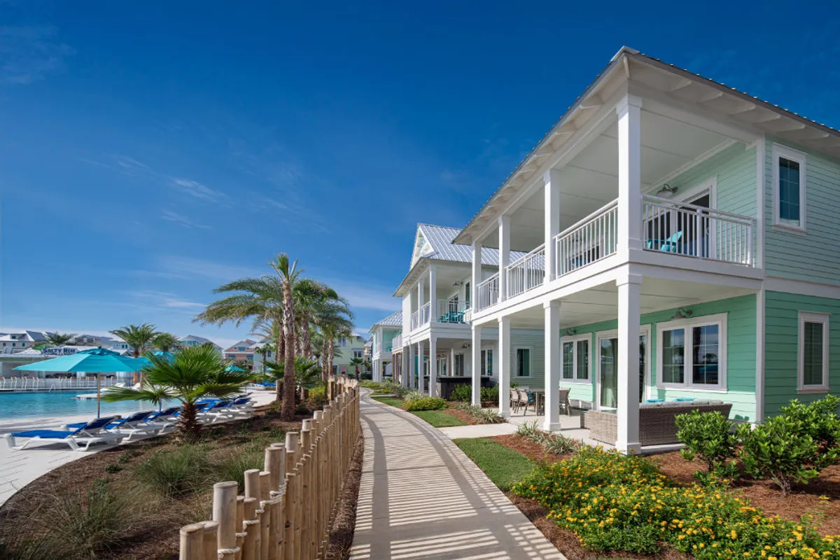 A vibrant exterior shot featuring turquoise and white beach cottages, each with balconies overlooking a pool area. Blue umbrellas and lounge chairs surround the pool, while a paved walkway lined with palm trees and lush greenery invites guests to explore. The scene is framed by a wooden fence that separates the walkway from the pool area, evoking a private, resort-like ambiance.