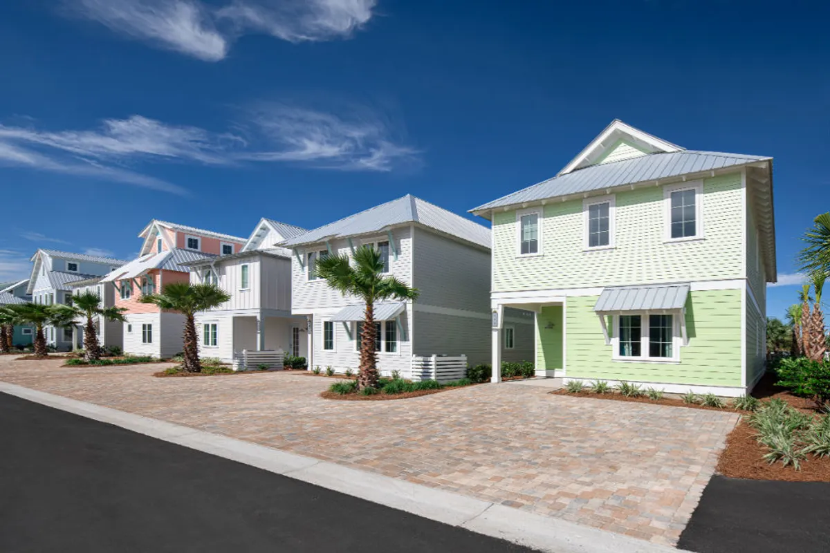 This image presents a row of colorful beach cottages with palm trees lining the driveways, well-paved stone driveways, and a bright blue sky. The scene captures the approach to the villas, with visible parking spaces right in front, and hints at the community’s inviting and well-maintained atmosphere.